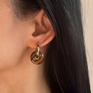 Può includere: Orecchini a cerchio dorati con un pendente in occhio di tigre. La pietra è di colore marrone e oro con un motivo a spirale.