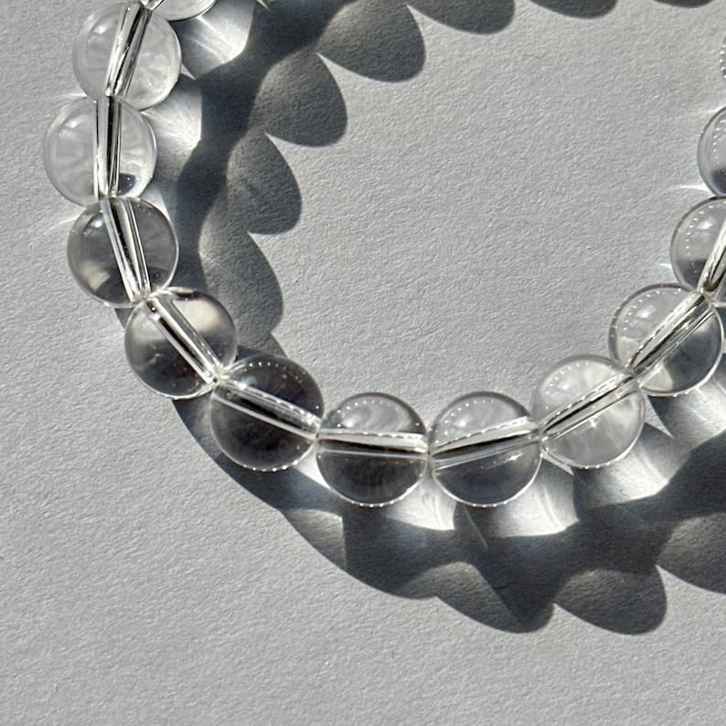 May include: A circular bracelet composed of smooth, spherical, transparent clear quartz beads strung together. The beads are polished to a high shine, reflecting light and casting distinct, scalloped shadows on the surface beneath the jewelry piece.