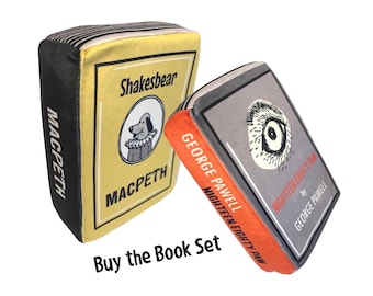 May include: Two plush book-shaped pillows. The first is yellow with black text that reads "MACPETH" and "Shakesbear" and features a cartoon dog. The second is gray with orange text that reads "NIGHTEEN EIGHTY PAM" and "GEORGE PAWELL".
