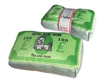 May include: A green plush toy shaped like a stack of money with the words "Royal Bone Bank" and "Miss Bone Digger" printed on it. The toy has a cartoon dog on it and says "100 £" on each side.