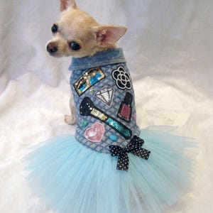 May include: A small tan Chihuahua dog wearing a denim vest adorned with sequined patches and a light blue tulle skirt. The vest features patches of a makeup brush, diamond, and flower. A black and white polka dot bow accents the skirt.