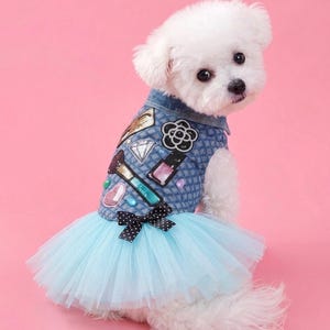 May include: A small white dog wearing a blue denim vest with a quilted texture and a light blue tulle skirt. The vest features sequined patches of a makeup brush, nail polish, a diamond, and a flower, finished with a black and white polka dot bow.
