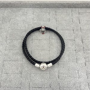 May include: Black braided leather bracelet with a silver charm featuring a detailed design and red accents. The bracelet has a double-wrap design and a silver clasp with the letters "PO".