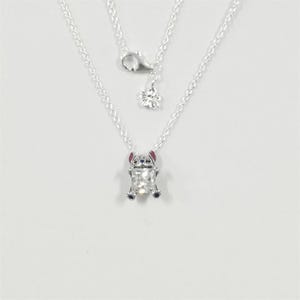May include: A silver necklace with a pendant depicting a cartoon character. The pendant is a clear, rectangular shape with a small, detailed figure of a character with pink ears and blue accents. The necklace has a delicate chain and a flower charm.