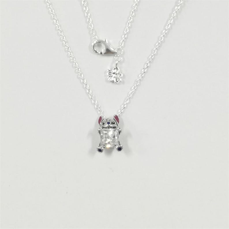 May include: A silver necklace with a pendant depicting a cartoon character. The pendant is a clear, rectangular shape with a small, detailed figure of a character with pink ears and blue accents. The necklace has a delicate chain and a flower charm.