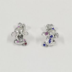 Puede incluir: Un par de pendientes de plata con un dise&ntilde;o de personaje de dibujos animados. Los pendientes tienen detalles de esmalte de colores en rosa, morado, azul y rojo. El personaje est&aacute; en una pose juguetona, con los brazos y las piernas extendidos.