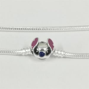 May include: A silver charm featuring a cartoon character's face with pink ears, black eyes, and a blue nose. The charm is attached to a silver chain, creating a bracelet or necklace component. The background is plain white.