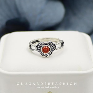 Pode incluir: Anel de prata com um design ornamentado com uma pedra vermelha redonda central. O anel tem uma banda detalhada e vazada e uma moldura decorativa ao redor da pedra. O texto "OLUGARDERFASHION Handcrafted Jewellery" está na parte inferior.