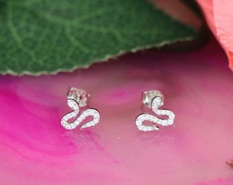 May include: A pair of silver snake earrings with clear cubic zirconia stones. The earrings are on a pink surface with a green leaf in the background.