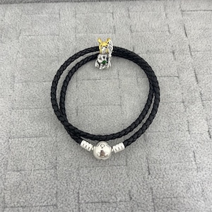 May include: Black leather wrap bracelet with a silver clasp and a charm featuring a gold-coloured figure with green accents. The bracelet is double-stranded and has a braided texture.
