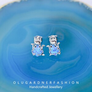 Könnte beinhalten: Silberne Ohrstecker mit einem blauen Opal-Design in Form einer kleinen Kreatur. Die Ohrringe sind vor einem blauen Hintergrund platziert. Der Text "OLUGARDNERFASHION Handcrafted Jewellery" ist unten im Bild zu sehen.