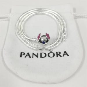 May include: A silver necklace with a charm featuring a cartoon character with pink ears, a blue nose, and black eyes. The necklace is coiled on a white pouch with the word "PANDORA" printed in black.
