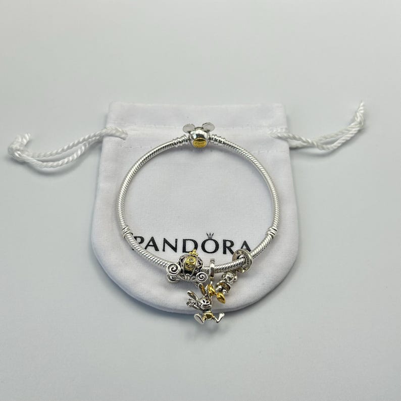 K&ouml;nnte beinhalten: Ein silbernes Pandora-Armband mit einem Mickey Mouse-Verschluss und -Anh&auml;nger. Das Armband hat einen goldfarbenen Mickey Mouse-Kopfverschluss und mehrere Anh&auml;nger, darunter eine goldfarbene Kutsche und einen Anh&auml;nger mit einer Zeichentrickfigur.