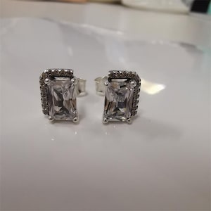 Peut inclure: Une paire de boucles d'oreilles à clous en argent. Chaque boucle d'oreille présente une pierre précieuse rectangulaire et transparente entourée d'un halo de petites pierres sombres. Les boucles d'oreilles sont posées sur une surface blanche.