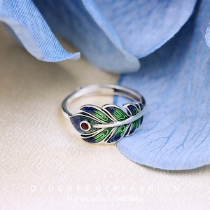 May include: A silver ring featuring a peacock feather design. The feather is green, blue and red with a red stone in the centre.