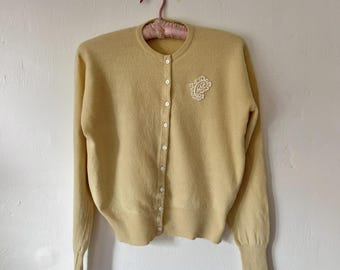 Puede incluir: Cárdigan amarillo pálido con mangas largas y cuello redondo. El suéter con botones presenta un diseño bordado blanco en el pecho izquierdo. El cárdigan está colgado de una percha acolchada rosa.