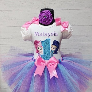 May include: A white tutu with a light blue and purple ombre effect. A white shirt with the text "Malaysia" and a glitter number "1" with two cartoon characters. The shirt has pink bows on the shoulders and a purple flower on the collar.