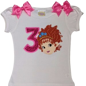 May include: White t-shirt with pink bows on the shoulders. Features a glittery pink number 3 and a cartoon character with red hair, a crown, and blue flowers. The shirt is designed for a child's birthday.