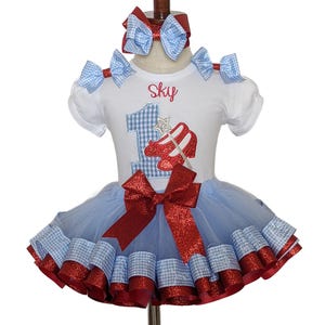 May include: A white short-sleeved dress with a blue tutu skirt, red accents, and a matching headband. The dress features a gingham "1" and ruby red slippers design, with the name "Sky" above. The outfit is designed for a first birthday celebration.