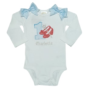 May include: White long-sleeved baby onesie with a blue gingham bow on each shoulder. The onesie has a blue number one with red ruby slippers and a star. The name Charlotte is embroidered below the number one.