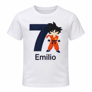 May include: A white short-sleeved t-shirt featuring a large dark blue number seven, a cartoon character with spiky black hair wearing an orange martial arts uniform, and the name Emilio printed in dark blue text below the graphic design.