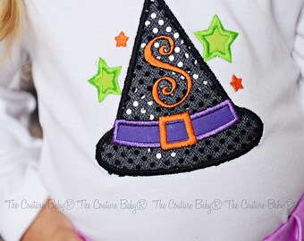 May include: White long-sleeved shirt featuring a black witch hat design with white polka dots, an orange letter "S", and a purple belt with an orange buckle. Green and orange star accents complete the Halloween theme. The shirt is paired with a purple skirt.