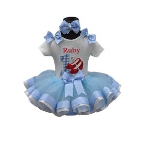 May include: A blue and white gingham tutu with a white shirt featuring a red number one and red ruby slippers. The shirt is personalized with the name "Ruby".