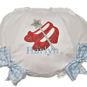 May include: White baby bloomers with blue gingham bows and a red embroidered image of ruby slippers and a magic wand. The name "Harlyn" is embroidered below the image.