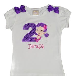 May include: White t-shirt with purple accents, featuring a mermaid design and the number "2" in sequins. The name "Teresa" is embroidered below the graphic. Two purple bows adorn the shoulders.