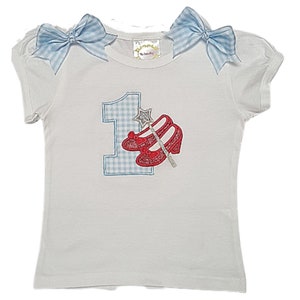 May include: White t-shirt with a blue gingham number one and red glitter ruby slippers with a silver wand. The shirt has blue gingham bows on the shoulders.
