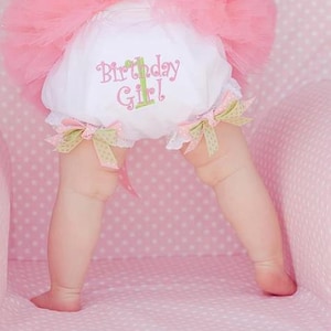 May include: A baby girl wearing a white tutu with a pink tulle skirt and a white onesie with the text "1 Birthday Girl!" embroidered on it.