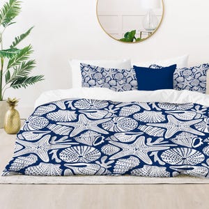 May include: A navy blue and white comforter set featuring a repeating pattern of starfish, seashells, and sea urchins. The set includes a comforter, two pillow shams, and a navy blue accent pillow. The bedding is displayed on a bed with a gold-framed mirror and a gold floor lamp.