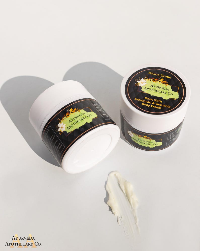 May include: Two white jars of body cream with black labels. The labels have green and gold accents and the text "Ayurveda Apothecary Co." and "Divine Drape Ghee With Sahatavari & Gotukola Body Cream". A dollop of white cream is on the table.