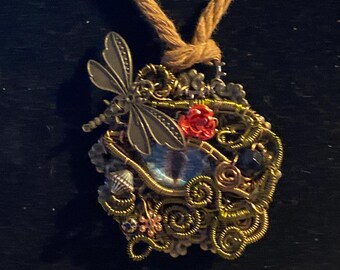 Puede incluir: Un collar dorado con un colgante que presenta una libélula, una flor roja y un ojo azul. El colgante está hecho de alambre y cuentas, y tiene una estética steampunk.