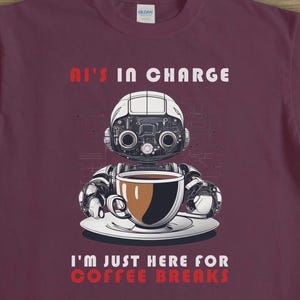 Op de afbeelding: Een maroon t-shirt met een witte robot die een kop koffie vasthoudt. De tekst op het shirt luidt "AI's in charge I'm just here for coffee breaks."
