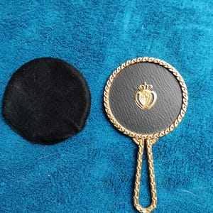 May include: A gold-colored hand mirror with a black leather backing and a gold crown and heart design. The mirror comes with a black velvet pouch.