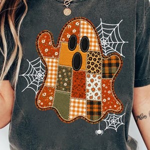 May include: A charcoal grey t-shirt featuring a large central ghost graphic composed of various patchwork patterns in orange, brown, and green tones. The ghost has three black oval cutouts for eyes and a mouth, surrounded by white spiderweb illustrations.