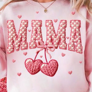 May include: Pink sweatshirt with the word "MAMA" in a woven design. The letters are white and pink. Below the word are two pink heart-shaped cherries tied with a bow. Small pink hearts are scattered around the design.