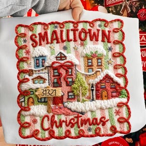Op de afbeelding: Wit geborduurd kunstwerk met een kerstthema met een kleine stadsscène met huizen, sneeuw en de woorden "Smalltown Christmas". Het ontwerp gebruikt rood, groen en wit, met het adres "31321" op een bord.