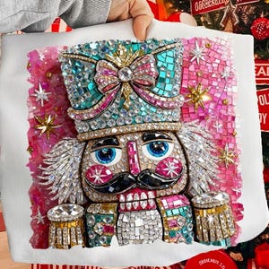 May include: A white tote bag with a mosaic design of a nutcracker soldier. The nutcracker has a teal and pink jeweled hat, blue eyes, and a black mustache. The design is surrounded by pink mosaic tiles and gold stars.