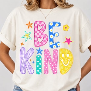 Op de afbeelding: Een cr&egrave;mekleurig T-shirt met korte mouwen en de tekst BE KIND in grote, kleurrijke, gedessineerde letters. Het ontwerp bevat roze ruitjes, blauwe bloemen, paarse zebramotieven, turquoise hartjes en gele smileys met diverse sterren.