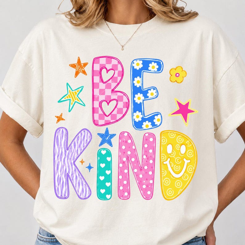 Op de afbeelding: Een cr&egrave;mekleurig T-shirt met korte mouwen en de tekst BE KIND in grote, kleurrijke, gedessineerde letters. Het ontwerp bevat roze ruitjes, blauwe bloemen, paarse zebramotieven, turquoise hartjes en gele smileys met diverse sterren.
