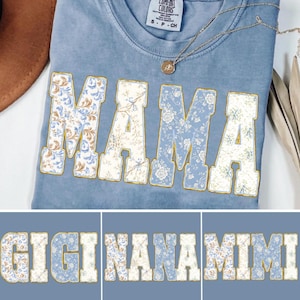 May include: A light blue t-shirt with the word "MAMA" in large, floral-patterned letters. The letters are outlined in gold. Below, the words "GIGI", "NANA", and "MIMI" are displayed in the same style.
