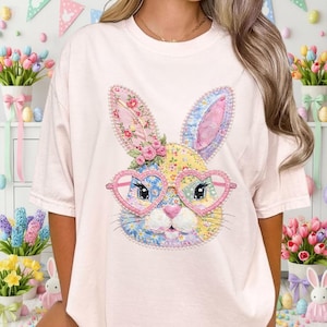 May include: A light pink t-shirt featuring a colorful patchwork bunny face with heart-shaped glasses. The bunny has pink, yellow, blue, and floral accents. The design is centered on the shirt, creating a whimsical and festive look.