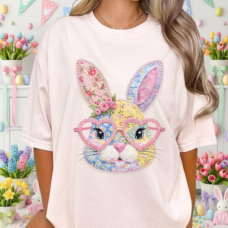May include: A light pink t-shirt featuring a colorful patchwork bunny face with heart-shaped glasses. The bunny has pink, yellow, blue, and floral accents. The design is centered on the shirt, creating a whimsical and festive look.