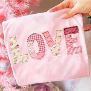May include: A light pink fabric with the word "LOVE" appliqued in various pink patterned fabrics. The letters are stitched onto the fabric, with the "L" featuring a floral pattern, the "O" a gingham check, and the "E" a striped pattern.