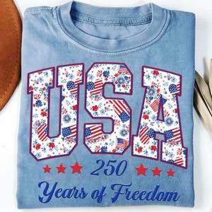 May include: Light blue t-shirt with the word "USA" in large, outlined letters filled with a floral and American flag pattern. Below the word "USA" are five red stars, the number "250", and the words "Years of Freedom" in blue.