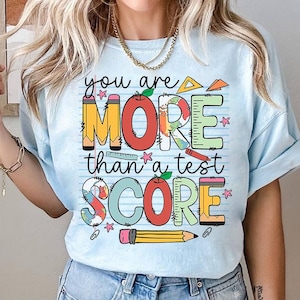 Peut inclure: Un t-shirt bleu clair avec un graphique coloré qui dit "You are more than a test score". Le graphique comprend un crayon, une règle, une pomme et un globe.