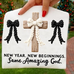 May include: A white sign with a wooden cross adorned with a cream-colored bow and two black bows. The sign reads "NEW YEAR. NEW BEGINNINGS. Same Amazing God."