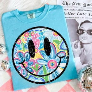 May include: A light blue t-shirt featuring a large smiley face design. The smiley face is filled with a floral pattern in shades of pink, blue, yellow, and green. The shirt is made of a soft material and is perfect for casual wear.